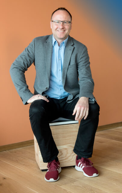 Axel Horstmann vor orangem Hintergrund auf Holzhocker
